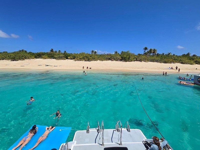 Looking from a boat's stern across turquoise water to a long sand beach, floating mats in the water
