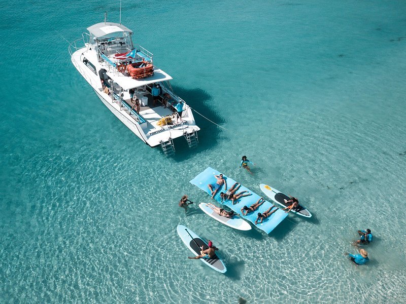 Aerial view of a catamaran with paddleboards and floating mats around it in turquoise water