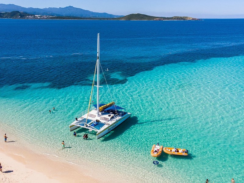 A sailing catamaran anchored off a white beach, a small inflatable beside it, seen from the air